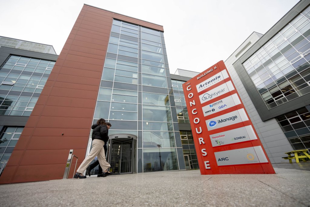 Modern office building with a red entrance tower and glass façade at The Concourse business park, Belfast. A large tenant directory sign featuring GEA and other companies stands in the foreground as two people walk towards the revolving entrance.